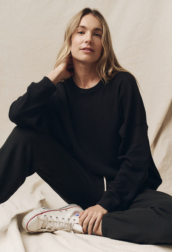 Person sitting on the floor wearing a black sweatshirt and pants, with white sneakers. Background is a neutral fabric.