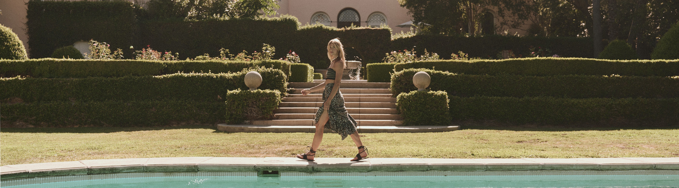 A woman in a patterned dress walks beside a swimming pool in front of a garden with trimmed hedges and a staircase.