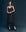 A woman stands against a dark background wearing The Legacy Dress - Black by THE GREAT. SP25 COLLECTORS, showcasing elegant fashion elements like a scarf around her neck, yellow socks, and black loafers.