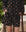 A person stands outdoors in THE GREAT. SP25 SLEEP The Square Pajama Short – Navy Cowboy, hands in pockets. A quilted blanket on the square and lush greenery add to the cozy, relaxed scene.