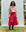 A person wearing a heart-patterned sweater, The Rhythm Skirt - Red Rose by THE GREAT. PS25, and brown boots stands outdoors against a light-colored backdrop.