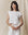 A person with long hair stands against a plain background, confidently wearing THE GREAT. SU23 CORE KNITS’ The Square Tee in True White along with white pants, looking at the camera.