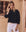 A person with long blonde hair in The Beaded Chevron Top by THE GREAT. SP25 D1 and white pants stands in front of a building with lattice windows, embodying the essence of women's fashion.