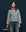 Wearing THE GREAT. SP25 COLLECTORS' The Slouchy Jean Jacket in Great Plains Wash, a person embodies a vintage-inspired look with matching jeans, standing beside a stool against a dark background.