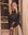 A person with long blonde hair slouches in front of a window, wearing THE GREAT. SP25 D1's The Western Slouch Sweatshirt in Washed Black and a beige skirt. They look directly at the camera, exuding a relaxed western charm.