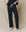 A person in The Wide Leg Cropped Sweatpant in Almost Black by THE GREAT. CORE KNITS stands against a beige background, lightly gripping the waistband of their pants, and wearing white sneakers.