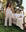 A woman wearing The Easy Sleep Pant in Cream Polka Dot by THE GREAT. PS25 SLEEP stands next to an outdoor metal bed with floral bedding, surrounded by trees and greenery, creating a serene backdrop for relaxation.