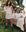 Wearing The Smocked Sleep Short in Cream Polka Dot by THE GREAT. PS25 SLEEP, a person stands next to an outdoor bed adorned with a teddy bear and floral pillows, capturing the cozy essence of loungewear amid serene trees and bushes.