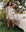 A person wearing The Nighttime Cami in Cream Polka Dot by THE GREAT. PS25 SLEEP stands beside an outdoor bed with pillows and a teddy bear, surrounded by grass, plants, and trees.