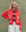 A woman stands against a green background wearing THE GREAT. SU25 The Bass Cardigan in Hot Red, featuring fish patterns, layered over a striped shirt and paired with a white skirt.