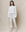 A person wearing THE GREAT. HOL 24 KNITS' The Slouch Sweatshirt in True White and matching sweatpants stands gracefully against a beige backdrop.