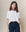 A woman in THE GREAT. SP25 KNITS' Short Sleeve Sweatshirt in True White, blue jeans, and a red patterned scarf stands against a neutral background. Her hands rest in her pockets, highlighting this perfect spring transition piece that combines comfort and style.