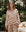 A woman stands outdoors wearing THE GREAT. SP25 SLEEP's The Pajama Shirt in Cream Cowboy, featuring a brown animal print; behind her, a teddy bear rests on a bench surrounded by greenery.