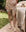 A person stands on a stone path in an outdoor garden square, wearing THE GREAT. SP25 SLEEP's The Square Pajama Short in Cream Cowboy, with a matching top and socks, beside a bed topped with a quilt and teddy bear.