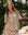 A woman wears THE GREAT. SP25 SLEEP's The Short Sleeve Pajama Shirt in Cream Cowboy, paired with matching shorts, standing outdoors by a bed adorned with pillows and a teddy bear amid lush greenery.