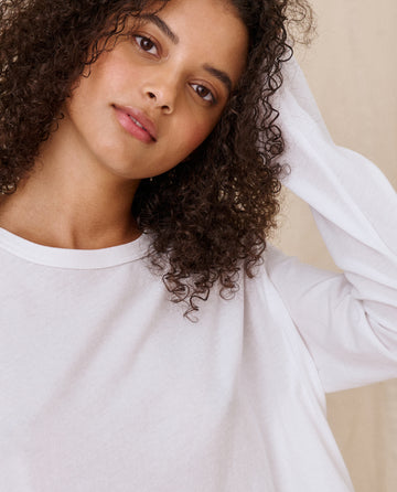 A person with curly hair wearing The Long Sleeve Crop Tee in TRUE WHITE by THE GREAT. CORE KNITS, looking slightly upward and touching their head.